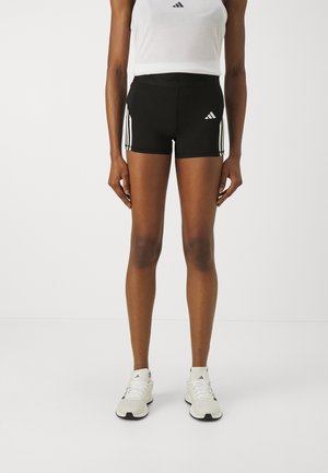 Persoon met zwarte Adidas-short met witte strepen en logo, witte sportschoenen en een wit mouwloos shirt tegen een effen achtergrond.