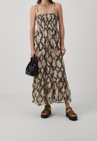 Nainen, jolla on yllään beige ja musta kuvioitu maksimekko ohuilla olkaimilla, pitää kädessään pientä mustaa käsilaukkua ja on pukeutunut ruskeisiin sandaaleihin.