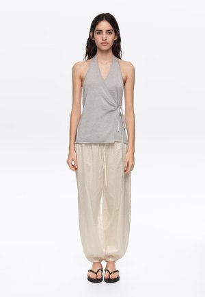 Mujer joven con un top halter plateado con un lazo lateral y pantalones sueltos de color beige claro, de pie y mirando hacia adelante sobre fondo blanco.