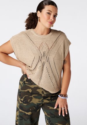 Femme aux cheveux bouclés portant un haut beige en tricot et un pantalon camouflage, avec des boucles d'oreilles anneaux et des bracelets, posant avec la main sur la hanche.