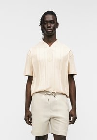 Μπεζ πουκάμισο με κοντά μανίκια, κάθετη ραβδωτή υφή και V-λαιμόκοψη. Συνδυασμένο με σορτς με κορδόνι στη μέση και διακριτικό μοτίβο.