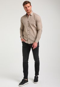 Camisa de botones en un beige claro con patrón geométrico; mangas largas, un bolsillo en el pecho. Jeans de mezclilla negros y zapatillas negras complementan el look.