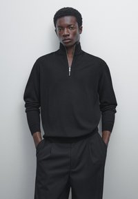 Black pullover with a quarter zip, long sleeves, and ribbed cuffs. Smooth texture, fitted silhouette, paired with tailored black trousers.
