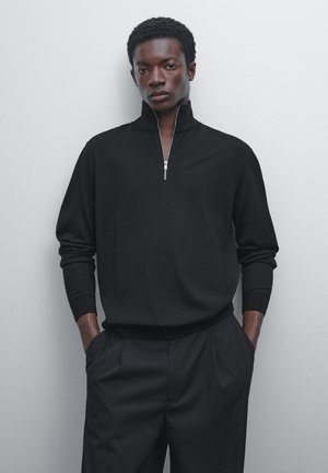 Black pullover with a quarter zip, long sleeves, and ribbed cuffs. Smooth texture, fitted silhouette, paired with tailored black trousers.