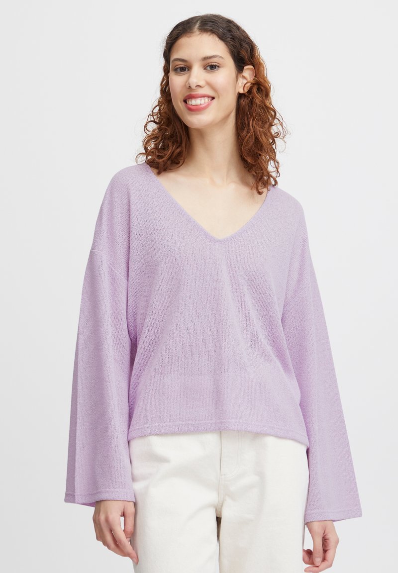 Pull violet clair en v avec des manches longues amples, confectionné en maille texturée, porté sur un pantalon blanc, mettant en avant sa coupe décontractée.