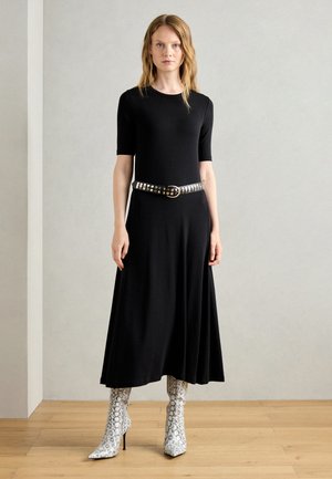 Robe noire, longueur genou, à manches courtes, accentuée par une ceinture noire cloutée. Associée à des bottines à talons aigus en imprimé serpent. Tissu lisse, design minimal.