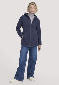 Marineblauwe ritsjack met capuchon, gemaakt van zachte stof. Beschikt over twee voorzakken. Draagt over een lichtpaarse coltrui en gecombineerd met een broek met hoge taille.