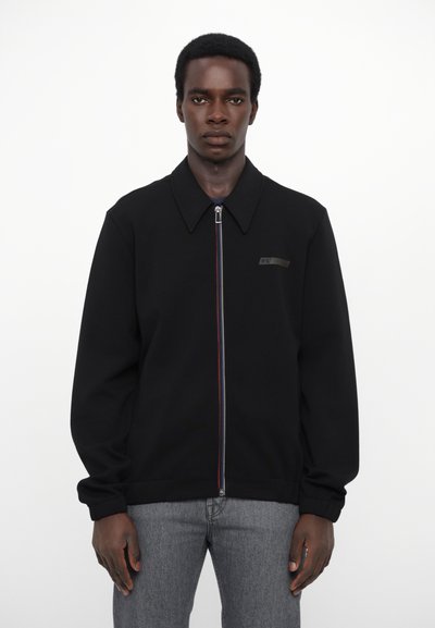 Schwarze Zip-Jacke mit Kragen, ausgestattet mit einem farbenfrohen Reißverschluss, dezentem Branding auf der Brust und einer lockeren Passform, gefertigt aus einem glatten Stoff.