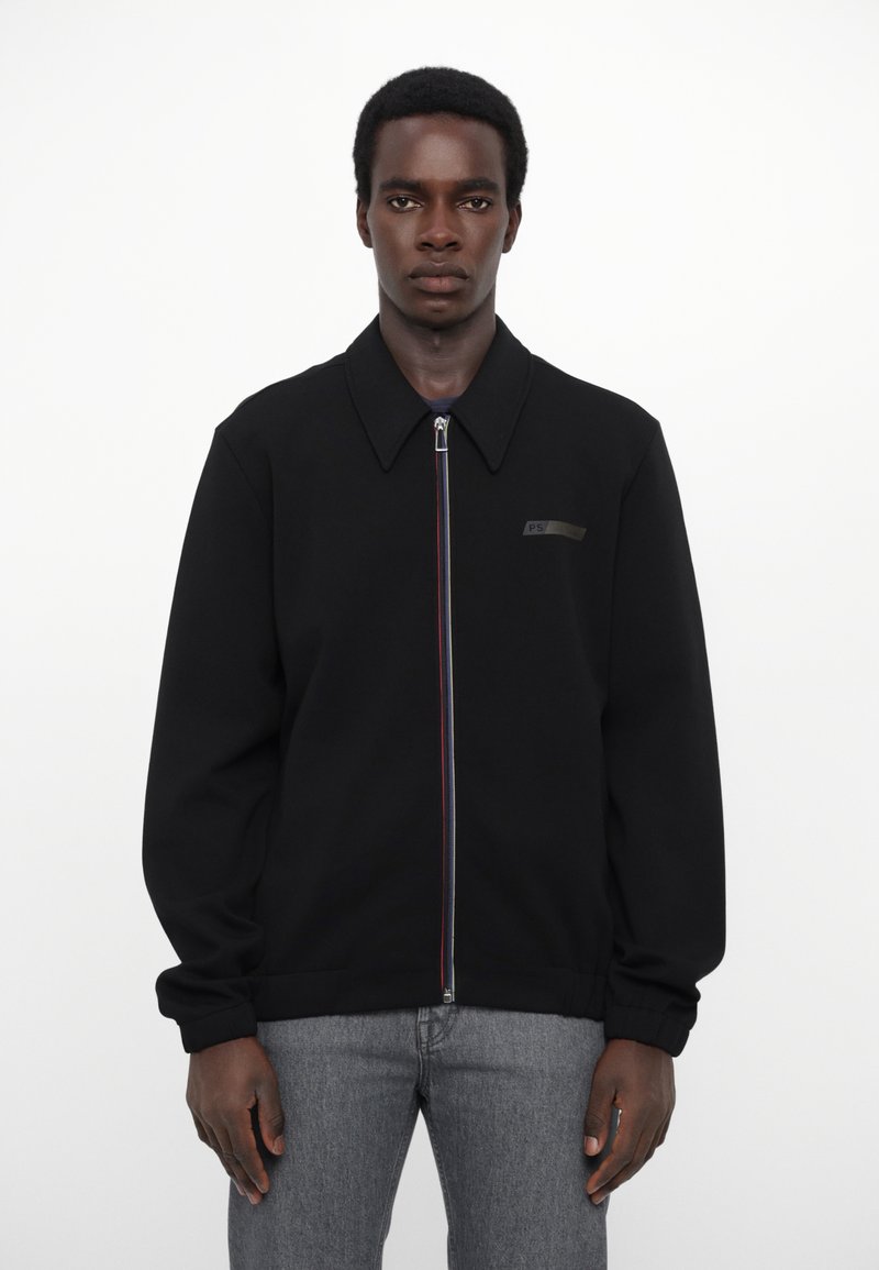 Veste zippée noire avec col, dotée d'une fermeture éclair colorée, d'un logo discret sur la poitrine et d'une coupe décontractée, fabriquée en tissu lisse.