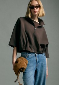 Woman with short blonde hair wearing black sunglasses, a dark zip-up cropped jacket, blue jeans, holding a brown suede bag against gray background.