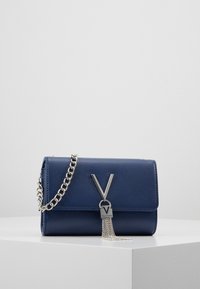 Sac à main en cuir marine avec une bandoulière en chaîne métallique, présentant un logo argenté et des franges. Forme rectangulaire avec une surface texturée.