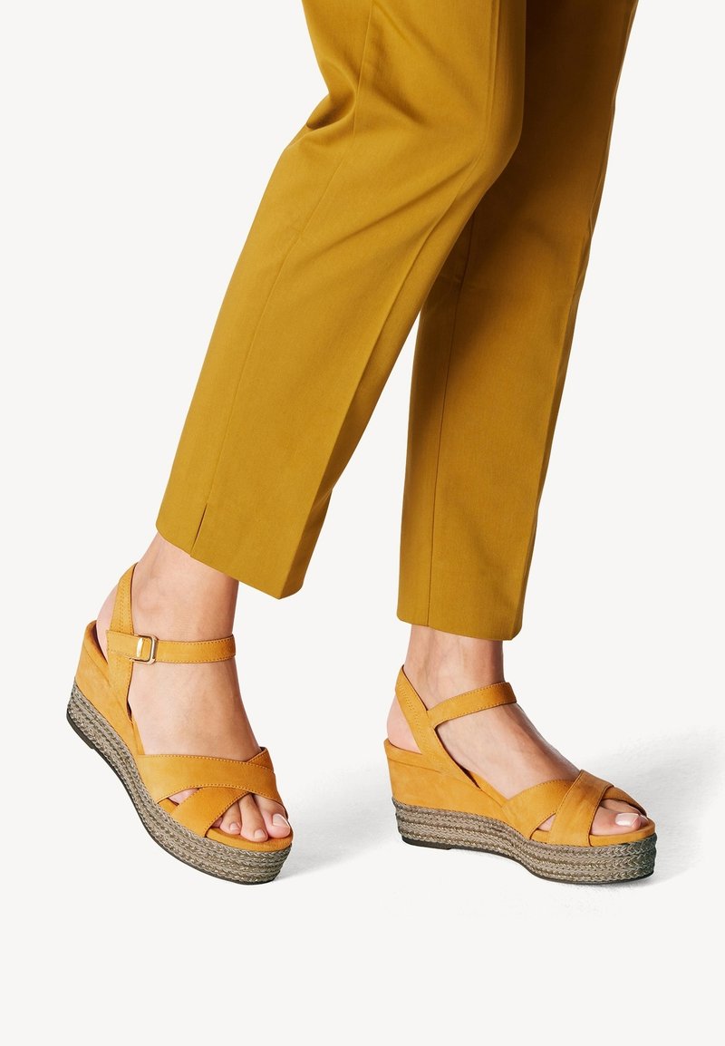 Sandalias de cuña de ante amarillo con tiras entrelazadas, puntera abierta y suela de yute trenzado, combinadas con pantalones ajustados color mostaza.