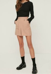 Mujer vestida con una blusa negra de manga larga, una falda mini beige con bolsillos delanteros y detalle de anillo, calcetas negras y mocasines negros, de pie contra una pared blanca.