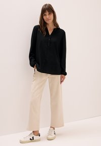 Schwarze Bluse mit langen Ärmeln, V-Ausschnitt und Knopfleiste, kombiniert mit beigen weitgeschnittenen Hosen und weißen Sneakers mit schwarzen Akzenten.