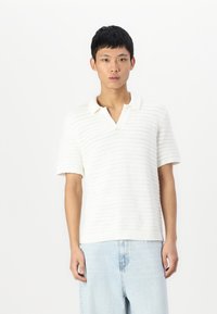 Camisa polo de punto blanca con un patrón texturizado, mangas cortas y cuello en V, combinada con jeans denim de color azul claro.