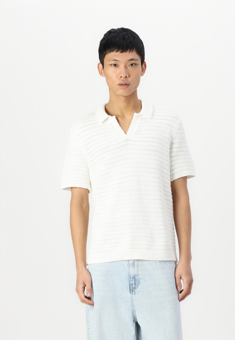Camisa polo de punto blanca con un patrón texturizado, mangas cortas y cuello en V, combinada con jeans denim de color azul claro.