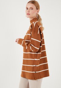 Maglione a righe marroni con righe orizzontali bianche, colletto alto, vestibilità oversize e bottoni decorativi sui lati. Texture liscia.