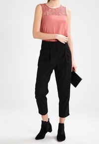 Haut sans manches rose clair avec détail en dentelle au niveau de l'encolure, associé à un pantalon noir taille haute et des bottines noires. Petite pochette simple à la main.