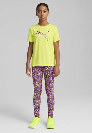 Neon-gelbes Sport-T-Shirt mit einem lila gemusterten Logo, kombiniert mit lila Leggings, die gelbe und schwarze abstrakte Designs aufweisen, sowie passenden Sneakers.