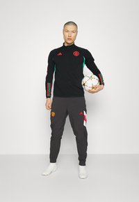 Top atlético negro con franjas verdes y rojas, logo del Manchester United. Pantalones grises con logo rojo y acentos blancos. El jugador sostiene un balón de fútbol.