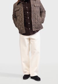 Brązowa kurtka w kratę z pikowanej tkaniny, z bocznymi kieszeniami, nałożona na ciemnobrązową koszulę z guzikami, w zestawieniu z białymi szerokimi spodniami i czarnymi butami.