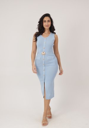 Kobieta w bezrękawowej, jasnoniebieskiej sukience midi z przodu zapinanej na guziki, z dwoma kieszeniami, ozdobną klamrą paska oraz przezroczystymi sandałami na wysokim obcasie.