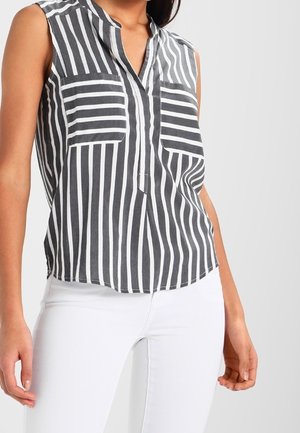 Blouse sans manches rayée verticalement noir et blanc avec deux poches poitrine à rayures horizontales, portée avec un pantalon blanc.