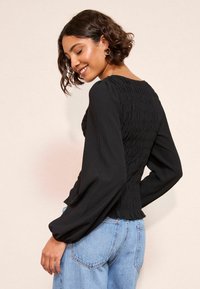 Młoda kobieta z krótkimi, kręconymi włosami uśmiecha się, nosząc złote kolczyki w kształcie kół, teksturowany czarny top z długim rękawem i jasnoniebieskie dżinsy, zwrócona lekko w tył.