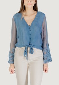 Blusa azul semitranslúcida con cuello en V, botones frontales y detalle de lazo. Las mangas largas tienen acentos bordados con lentejuelas. Combinada con pantalones claros.