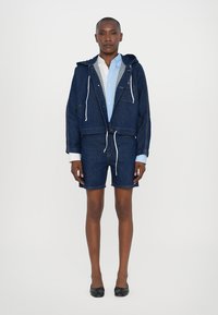 Denimjakke med hætte og snøringer, parret med matchende denimshorts. Lyseblå skjorte underneath, med en let tekstureret stof.