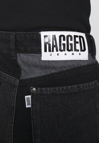 Jeans in denim nero con un grande etichetta bianca recante la scritta "RAGGED JEANS". Presentano una distintiva macchia scura sulla vita e una piccola etichetta sulla tasca.