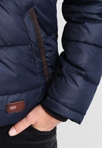 Chaqueta de puff navy con detalles en cremallera marrón, patrón acolchado y un parche de cuero etiquetado "1977" en el lado inferior. Textura suave, diseño cálido.