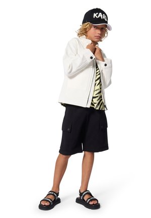 Veste en jean blanche, shorts cargo noirs, sandales noires et blanches avec logo, chemise verte et noire à motifs, casquette noire avec lettrage blanc.