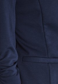 Detail rukávu tmavě modrého saka se zapínáním a postranní kapsou, ukazující strukturu látky a precizní šití.