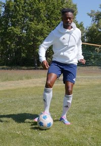 Hvit hettegenser med snorer, blå shorts, hvite sokker med svarte striper, og flerfargede fotballsko, som dribler en mønstret fotball på gress.