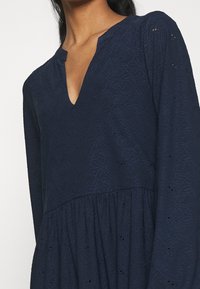 Robe bleu marine avec un décolleté en V, des manches longues et un motif en dentelle florale. Présente une taille froncée et un tissu textur é.