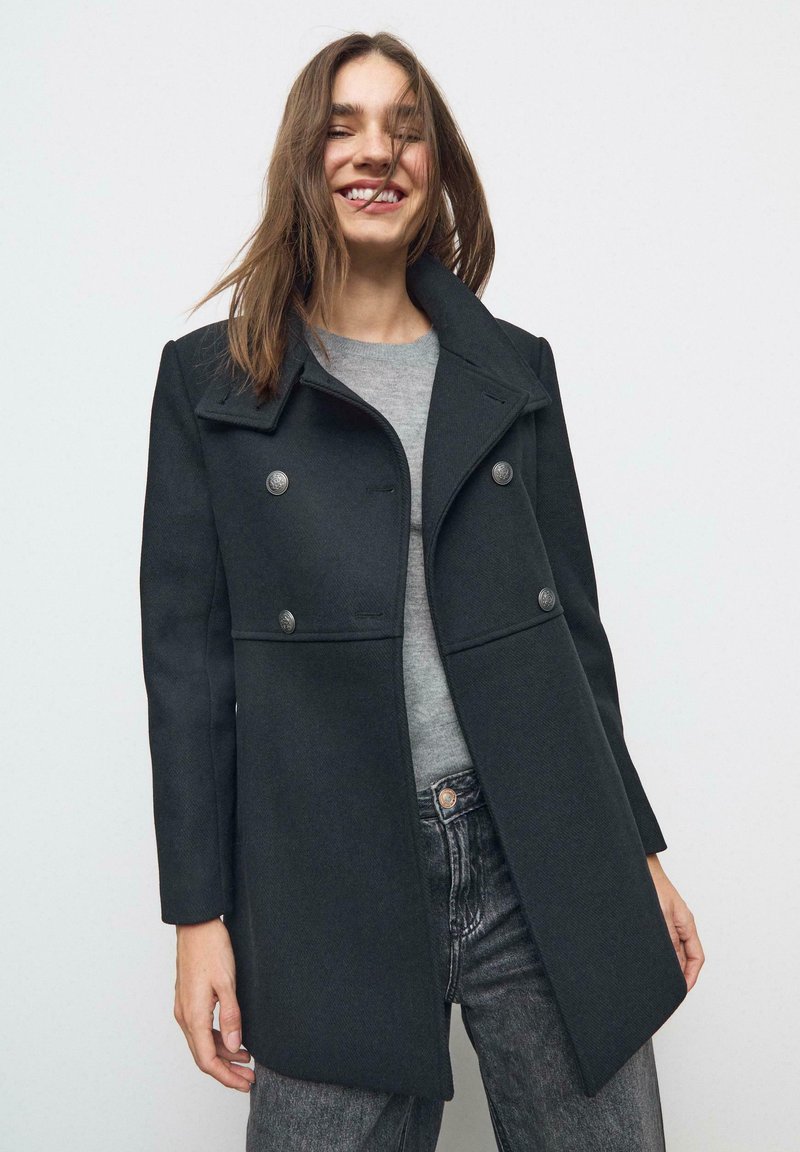 Cappotto doppiopetto in misto lana nero con colletto a ragged, bottoni argentati e texture liscia, indossato sopra un top grigio e jeans chiari.