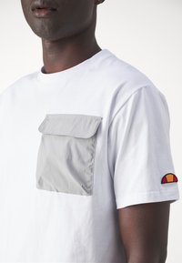 Hvid bomulds t-shirt med en grå klaplomme, korte ærmer og et farverigt emblem på venstre ærme. Glat tekstur.