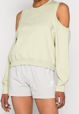Femme portant un haut à manches longues vert clair avec épaules dénudées et un short gris clair, tous deux avec des logos Converse visibles.