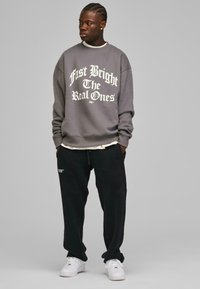 Grå sweatshirt med hvit tekst, avslappet passform. Svart joggebukse, løs struktur, med sidelommer. Hvite sneakers med kraftige såler.
