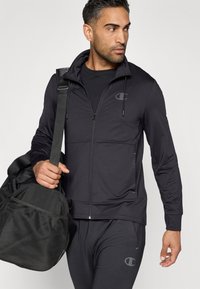 Schwarze Zip-Up Kapuzenjacke mit taillierter Passform, aus glattem Stoff, mit einem Logo auf der Brust und einer Reißverschlusstasche, kombiniert mit einer schwarzen Sporttasche.