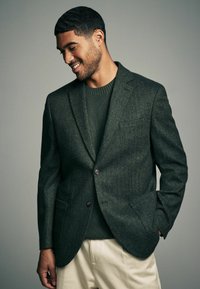 Dunkelgrüner Blazer mit Fischgrätmuster und zwei Knöpfen, getragen über einem passenden Strickpullover. Cremefarbene Hosen vervollständigen das Outfit.
