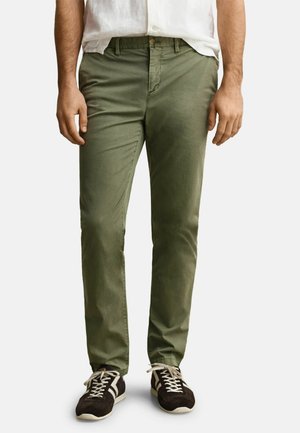 Homme portant un pantalon slim vert olive et des baskets noires avec des lacets blancs, debout devant un fond uni.