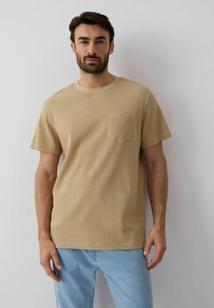 Mężczyzna o ciemnych włosach i brodzie, ubrany w beżowy krótki teksturowany t-shirt z kieszenią na piersi i jasnoniebieskie dżinsy, stojący na tle gładkiej ściany.