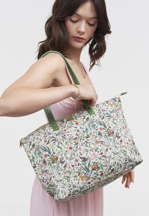 Woman in pink dress holding large floral tote bag with green handles featuring various animal and plant illustrations.