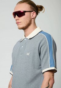 Polo à rayures bleu et blanc, avec un col et des accents de manches contrastants. Le mannequin porte des lunettes de soleil élégantes et teintées.