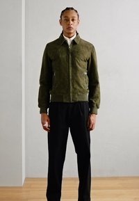 Giacca bomber in camoscio verde oliva con zip frontale, tasche laterali e polsini a costine, abbinata a pantaloni neri. Tessuto morbido e design aderente.