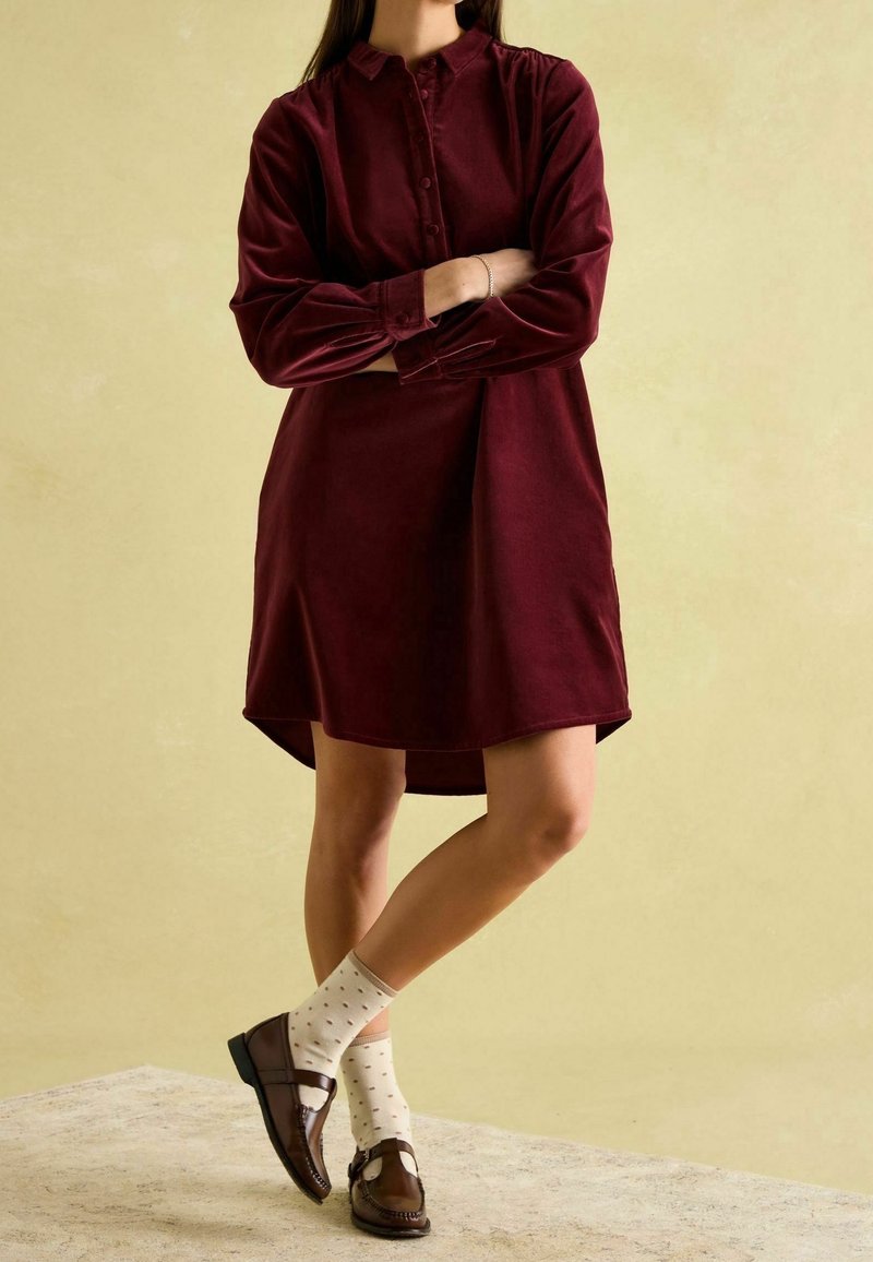 Personne debout, bras croisés, portant une robe bordeaux à manches longues, des chaussettes crème à pois et des mocassins marron foncé sur un fond uni.