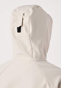 Svetlo bež jakna s kapuco in majhno črno zanko na vrhu kape, prikazana od zadaj.