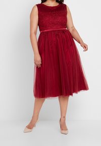 Bordeaux jurk met een mouwloze, geborduurde lijfje, een tule rok en een tailleband; gecombineerd met beige hoge hakken. Gladde stof textuur.